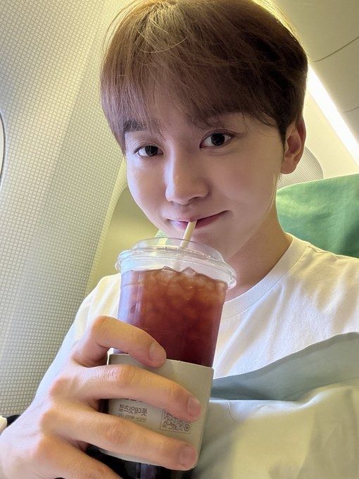 a young man holding a drink in his right hand and looking at the camera while sitting on an airplane