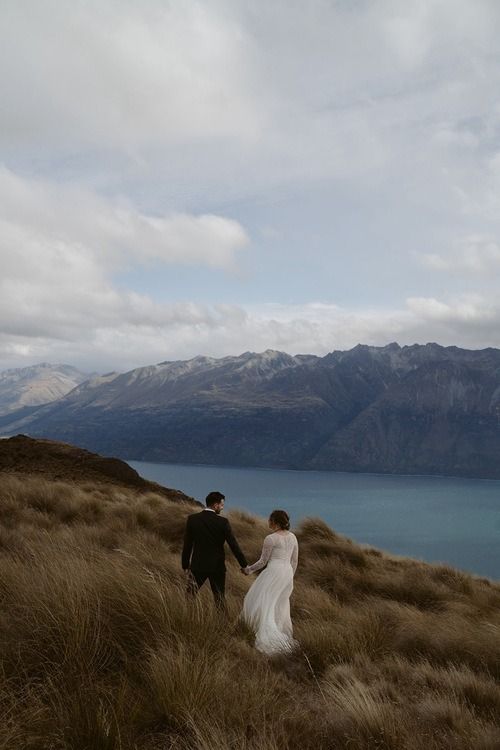 a bride and groom holding hands while walking up a hill near the water with mountains in the background