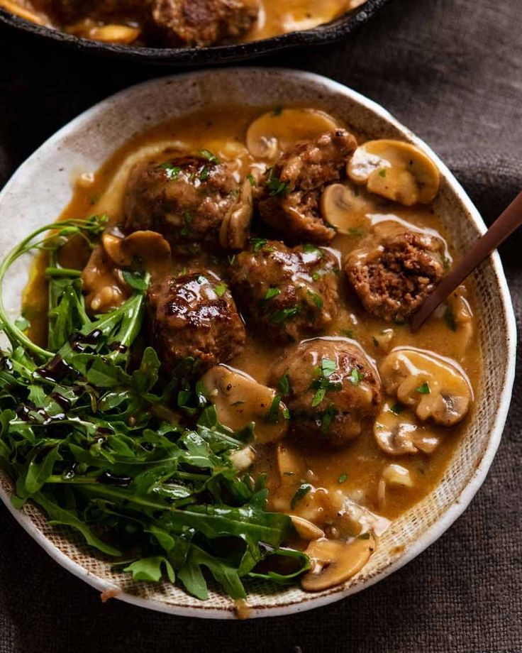 two bowls filled with meatballs and mushrooms covered in gravy next to greens