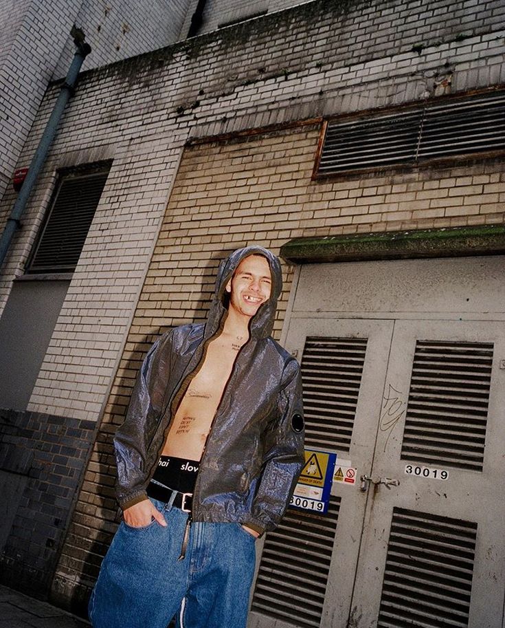 a young man standing in front of a brick building with his shirt off and hood up