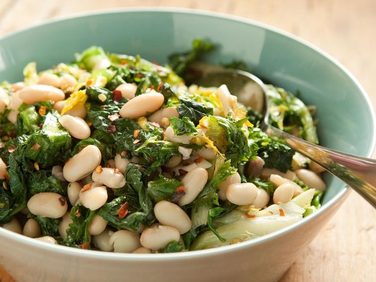 a bowl filled with white beans and greens on top of a wooden table next to a spoon