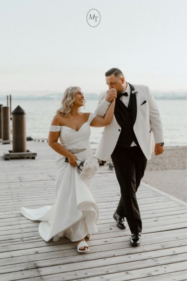 a bride and groom walking on a dock near the ocean holding each other's hands