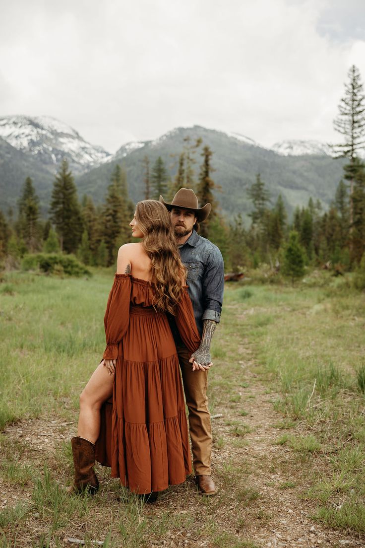 a man and woman standing next to each other in the grass with mountains behind them