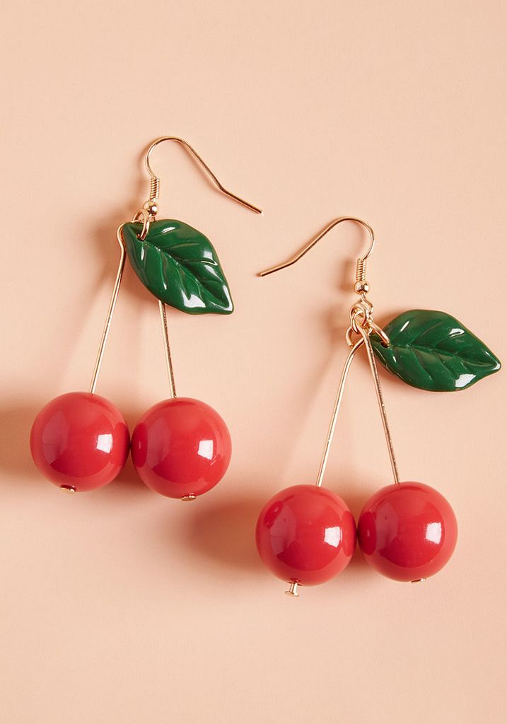 three cherries with green leaves hang from gold earwires on a pink background