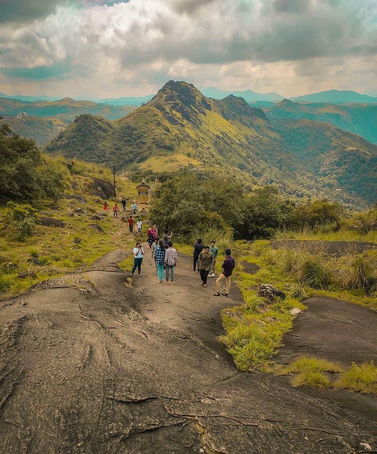 a group of people walking down a dirt road in the middle of mountains with green grass on both sides