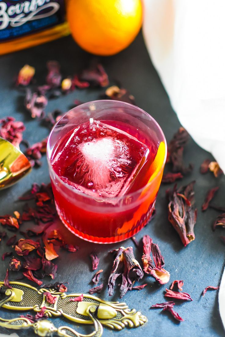 a glass filled with red liquid next to an orange and some dried flowers on a table