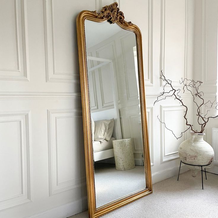 a large mirror sitting on the floor in front of a white wall next to a vase with flowers