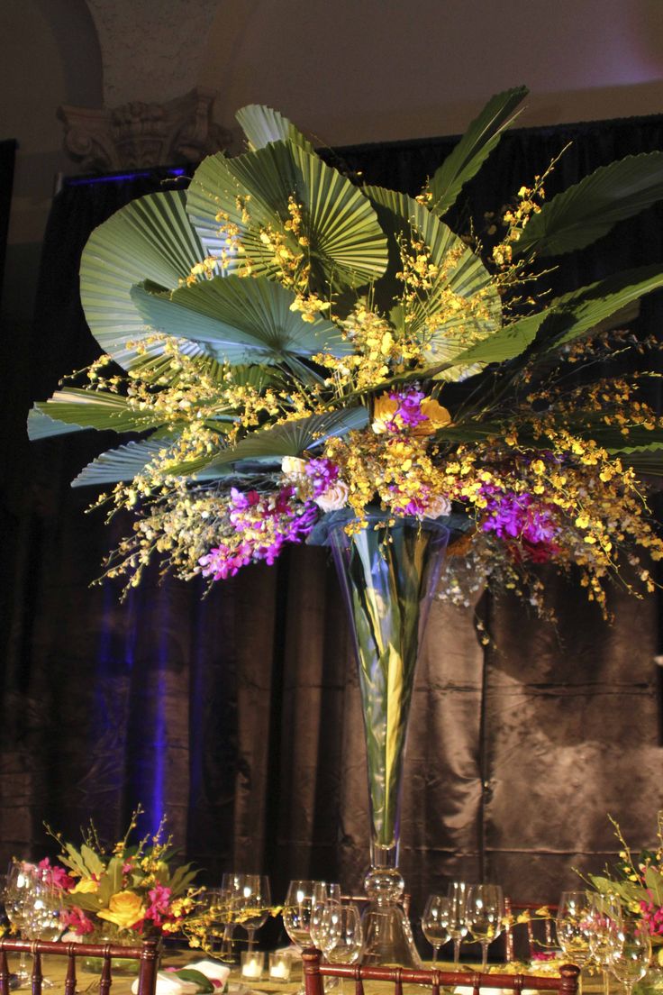 a tall vase filled with flowers and greenery on top of a table covered in wine glasses