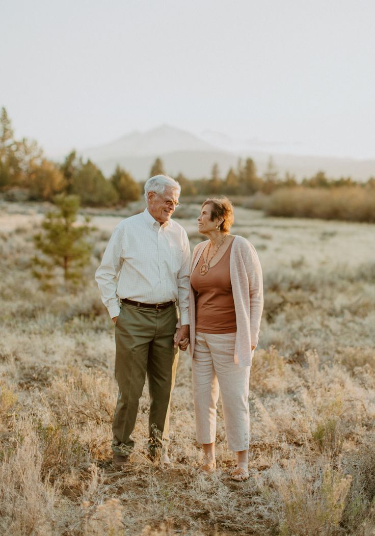 an older man and woman holding hands in the middle of a field with mountains in the background