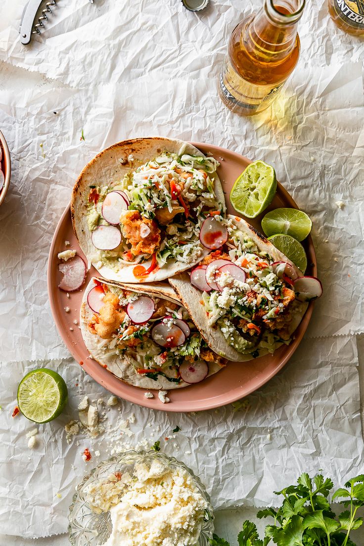 two tacos on a pink plate with limes, cilantro and other ingredients