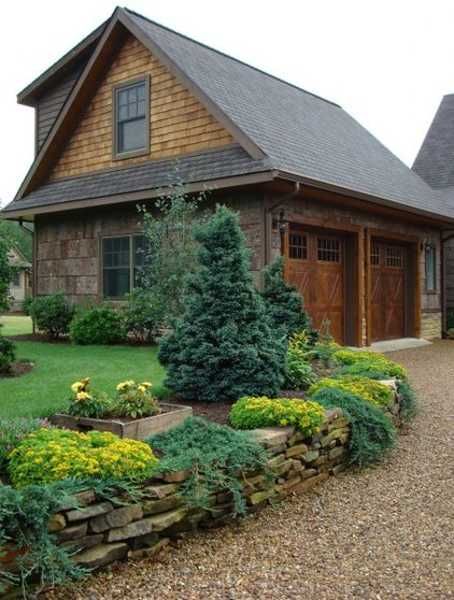 a stone wall with plants and flowers in front of a barn style house on a gravel driveway