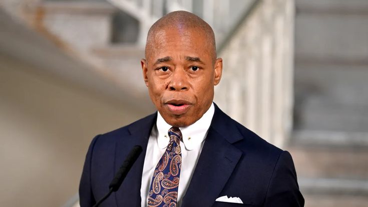 a man wearing a suit and tie standing in front of some stairs with his hand on his hip