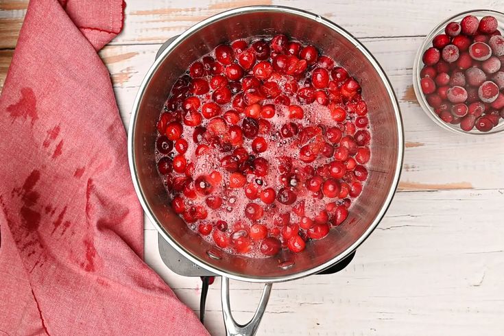 a pot filled with water and cranberries on top of a wooden table next to a red cloth