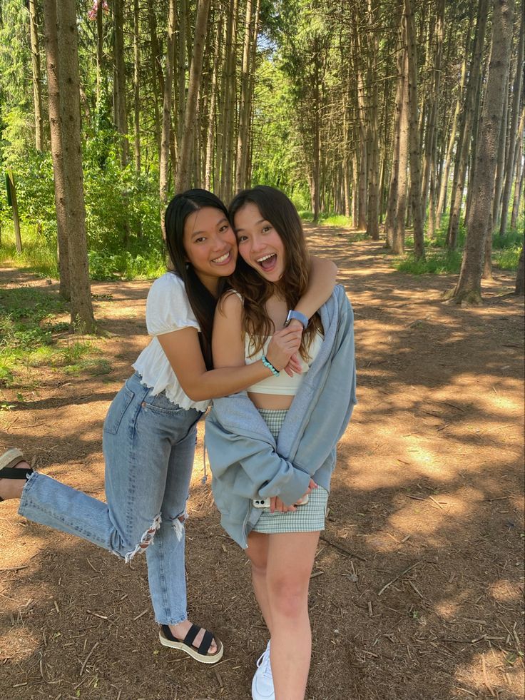 two young women hugging each other in the middle of a wooded area with trees behind them