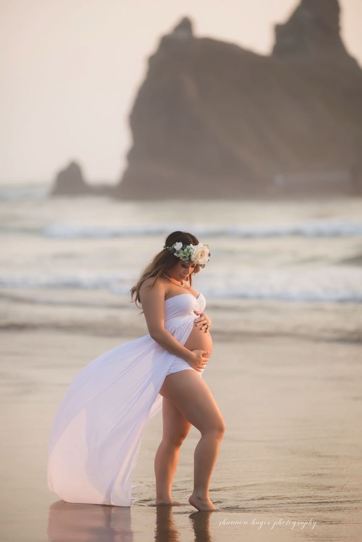 a pregnant woman in a white dress standing on the beach with her arms around her waist