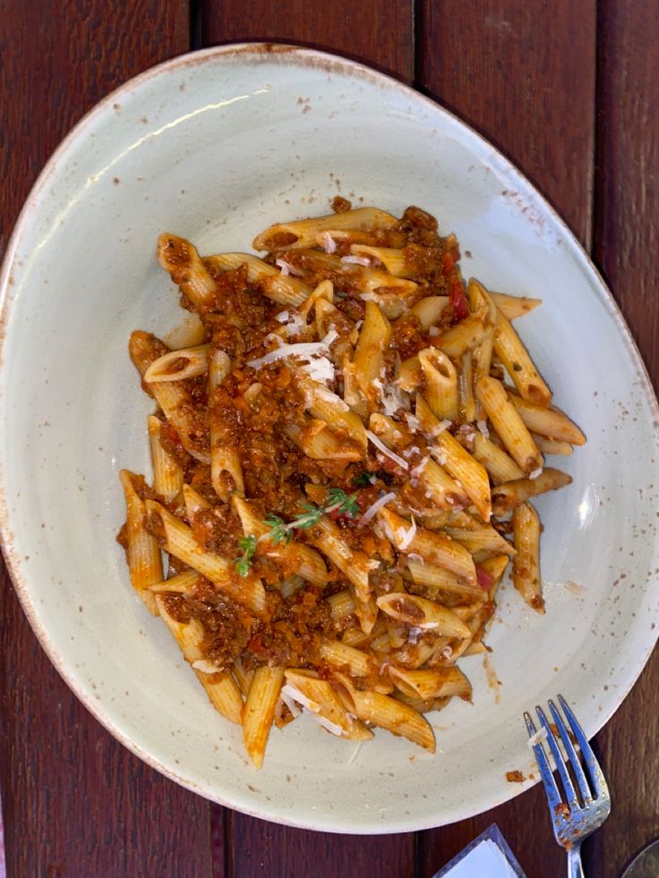 a white bowl filled with pasta and sauce on top of a wooden table next to utensils