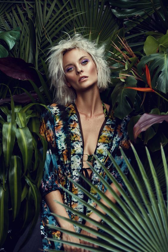 a woman with white hair is posing in front of some tropical plants and palm leaves