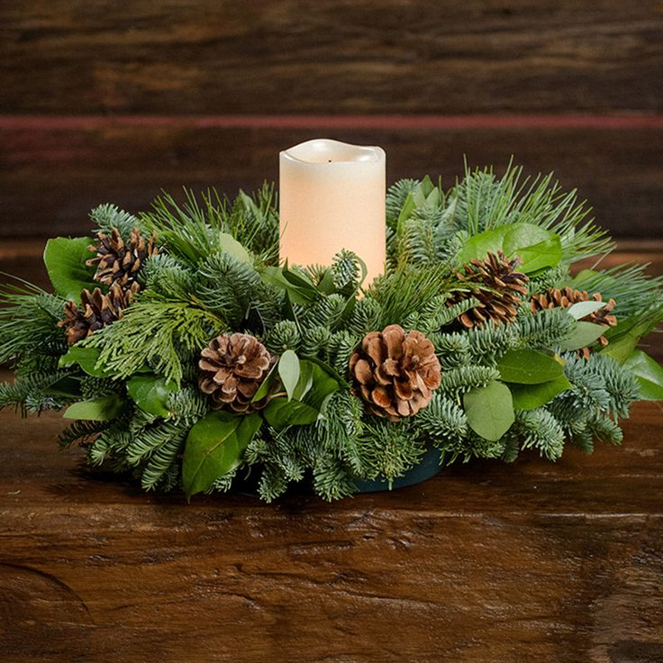 An arrangement of noble fir, cedar, white pine, salal, bay leaves, pinecones, and an ivory LED candle sitting on a wooden shelf with a dark wood background. Christmas Lantern Centerpieces, Salal Leaves, Cranberry Centerpiece, Nautical Centerpiece, Candle Decorations, Ivory Candles, Star Candle Holder, Red Napkins, Christmas Dining Table