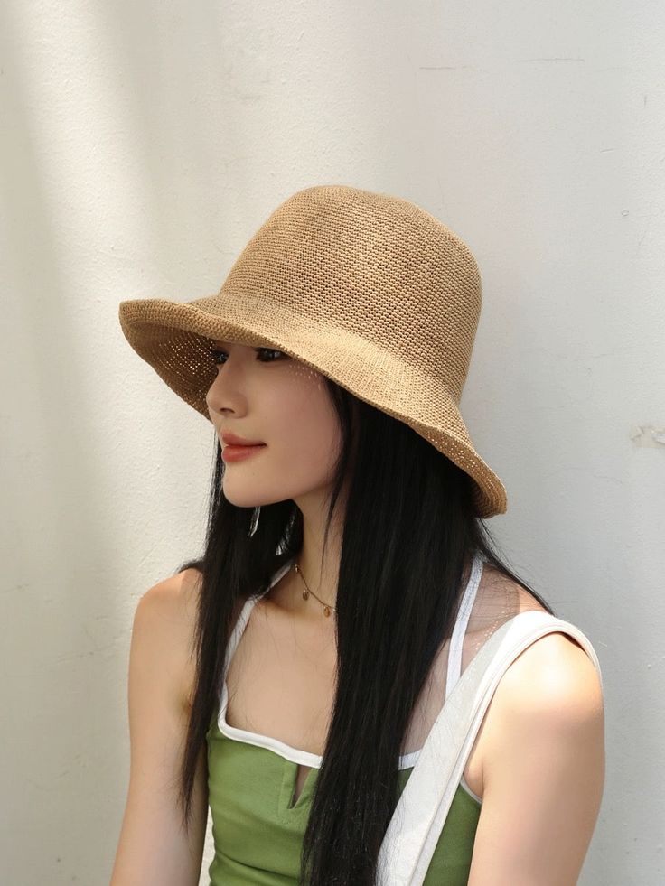 a woman with long black hair wearing a tan hat and green tank top standing against a white wall