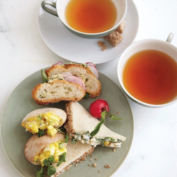 two plates with sandwiches and cups of tea next to each other on a white table