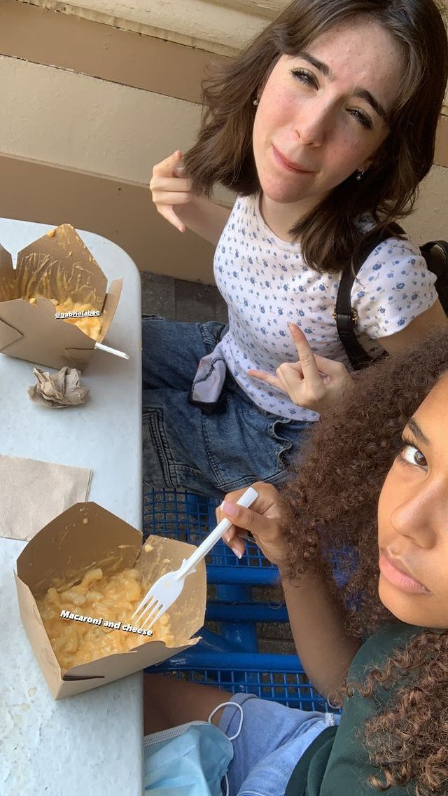 two women sitting at a table with food in boxes on the table and one holding a fork