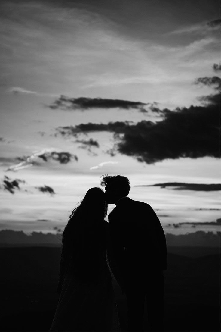 black and white photo of two people kissing in front of the sky with clouds behind them