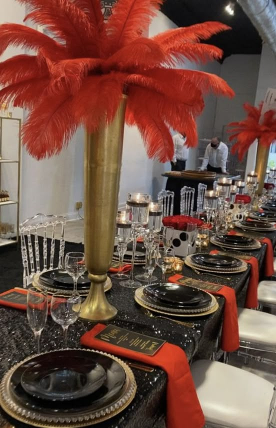 a table set up with black and gold plates, silverware and red feather centerpieces