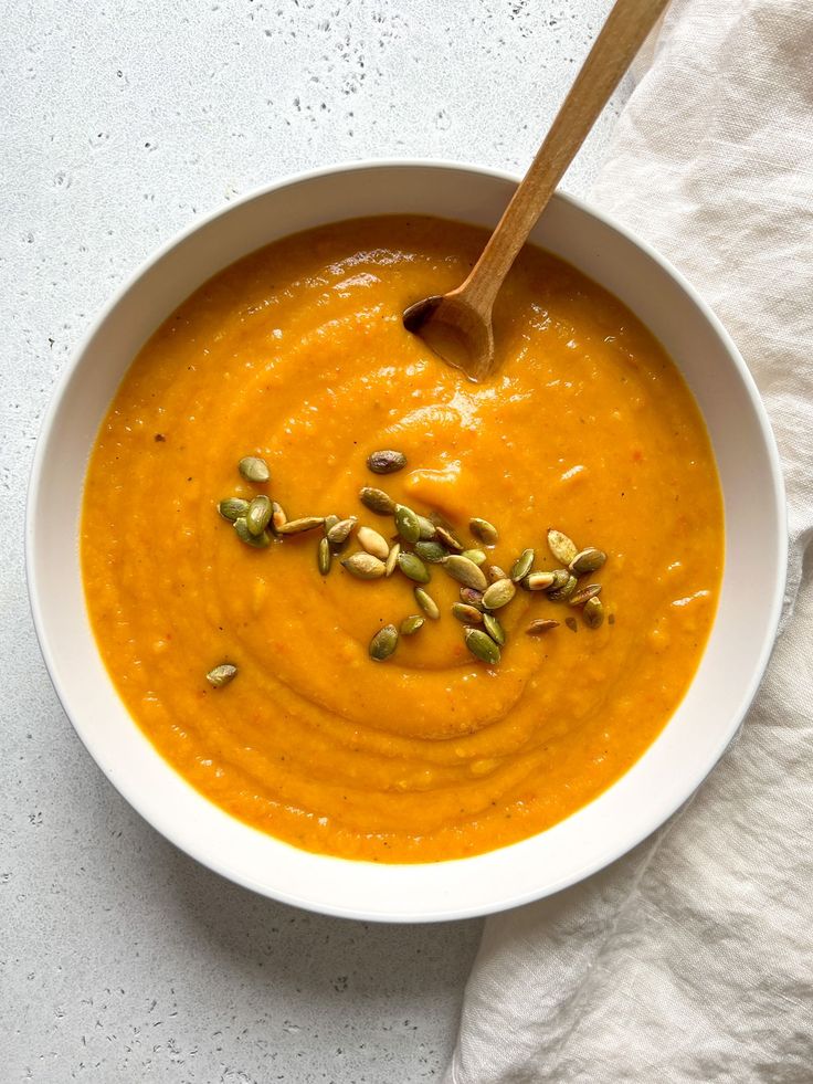 a white bowl filled with carrot soup and topped with pumpkin seeds next to a wooden spoon