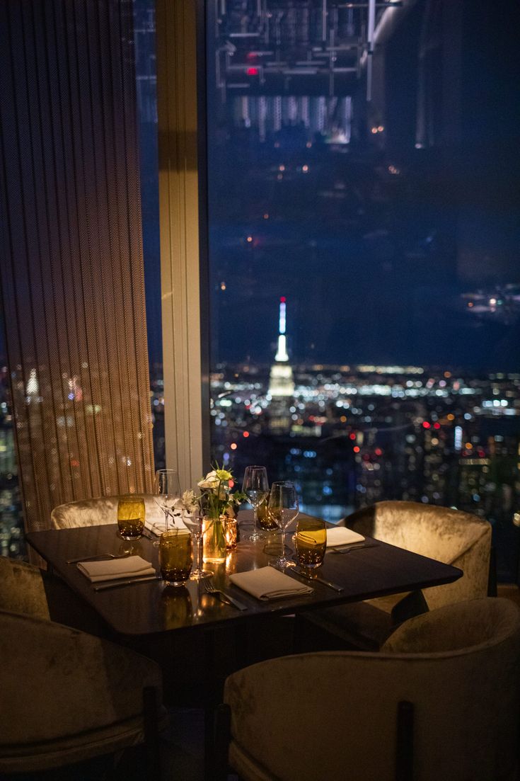 a table with glasses and plates on it in front of a window overlooking the city at night