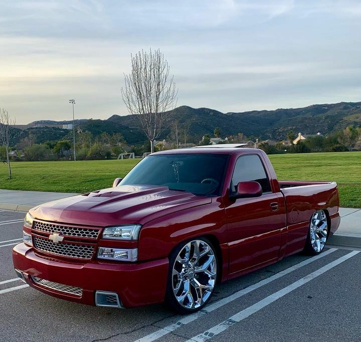 a red pick up truck parked in a parking lot next to a grassy area with mountains in the background