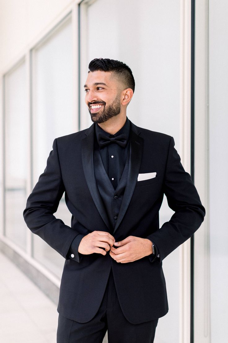 a man in a black tuxedo smiles while standing next to a white wall