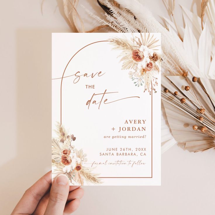 a person holding up a wedding card with flowers and feathers around it on top of a table