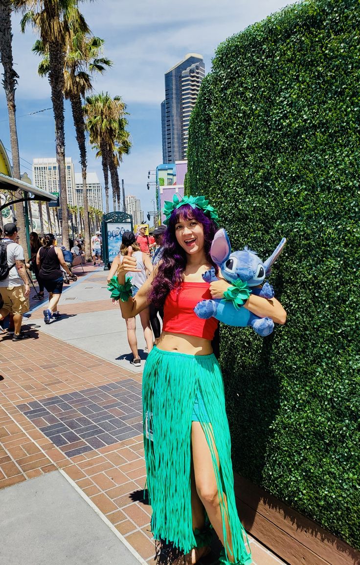 a woman in a green skirt and red top standing next to a tall plant wall