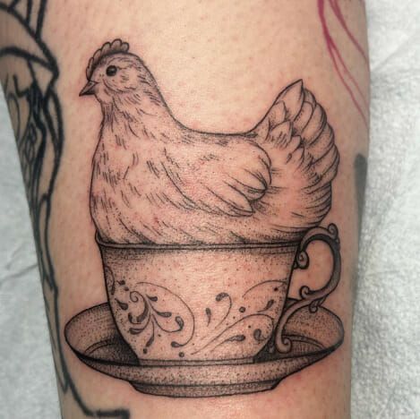 a chicken sitting in a teacup on the leg with tattoos around its neck and arm