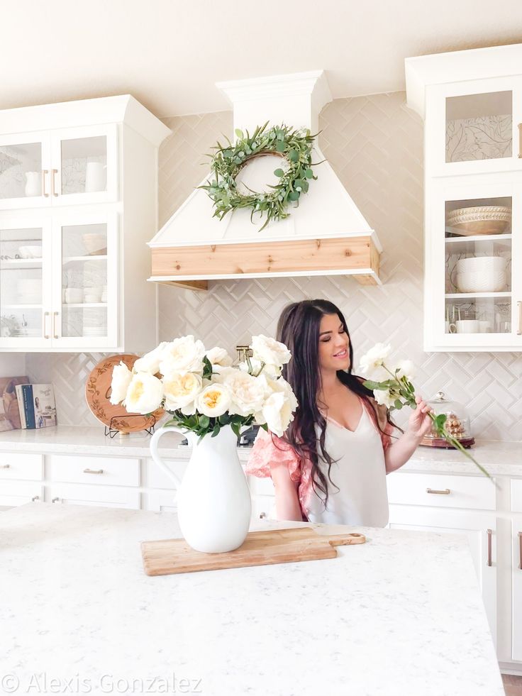 a woman standing in a kitchen with flowers on the counter and white cabinets behind her