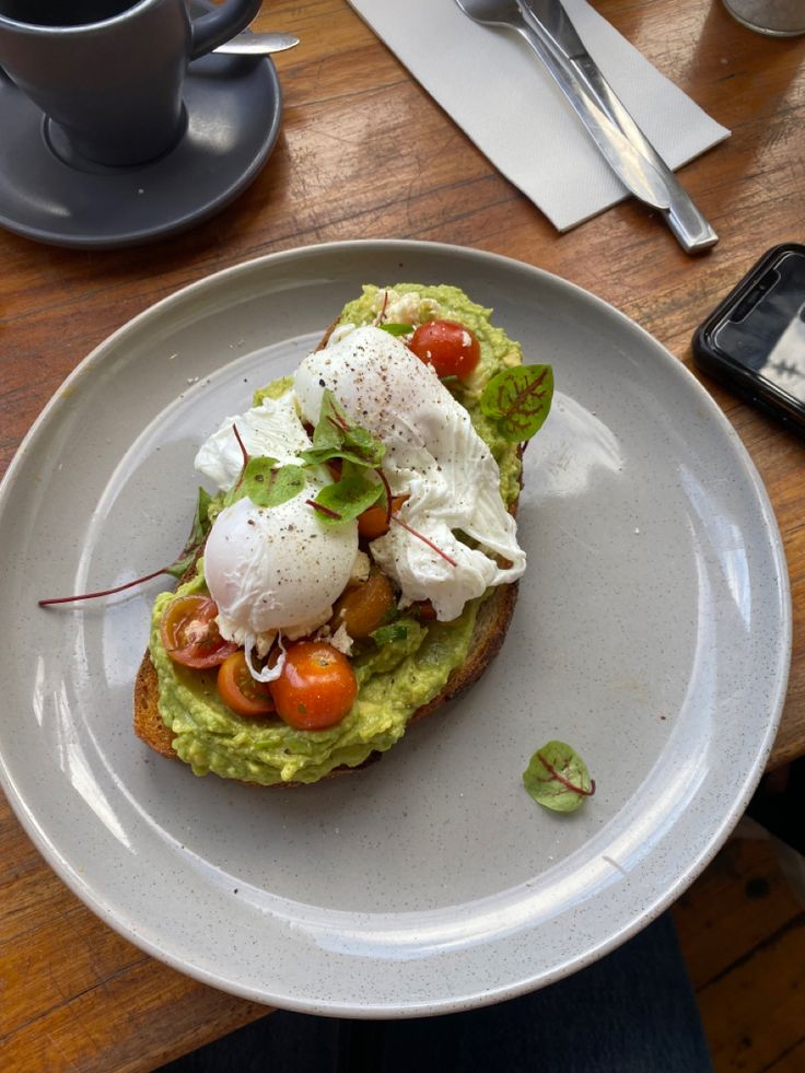 an avocado toast topped with poached eggs and cherry tomatoes sits on a plate next to a cup of coffee