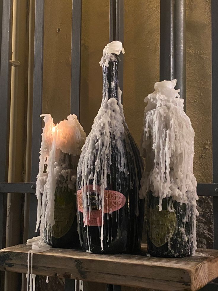 three wine bottles covered in ice on a wooden shelf next to a metal fence with candles