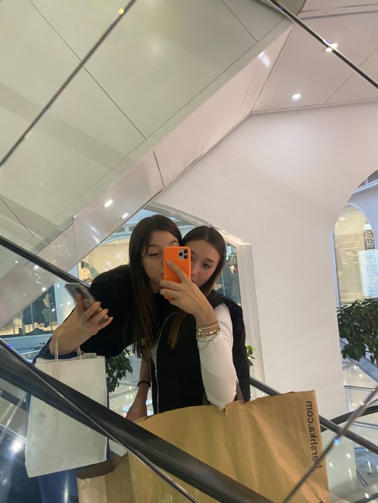 two women taking pictures on their cell phones while walking down the escalator with shopping bags