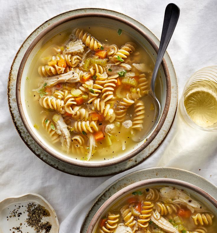 two bowls filled with chicken noodle soup next to a glass of wine and spoon