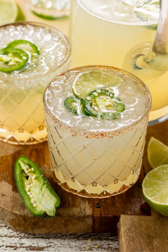 two margaritas with lime and jalapenos sit on a wooden cutting board