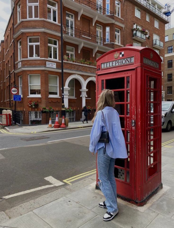 a woman standing next to a red phone booth on the side of a road with buildings in the background