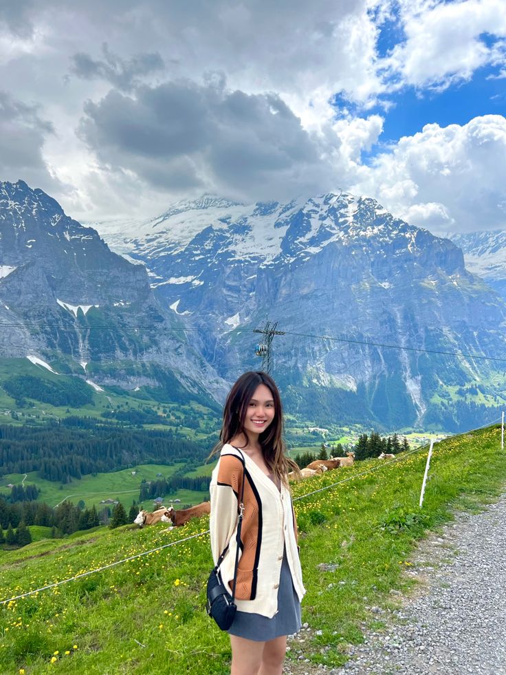 a woman standing on the side of a road in front of mountains and grass covered hills