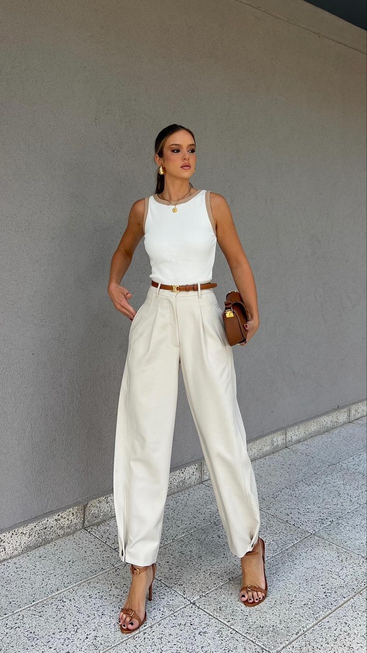 a woman standing in front of a gray wall wearing white pants and a white top