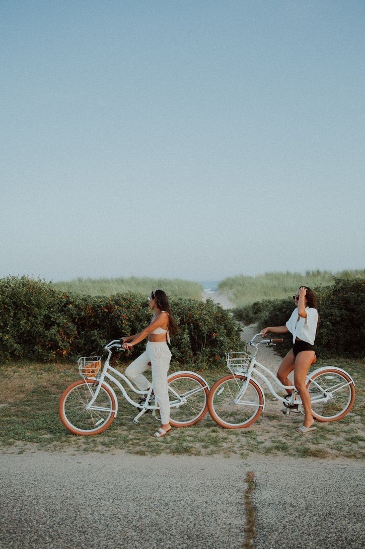 two women riding bikes on the side of a road next to grass and bushes with orange rims