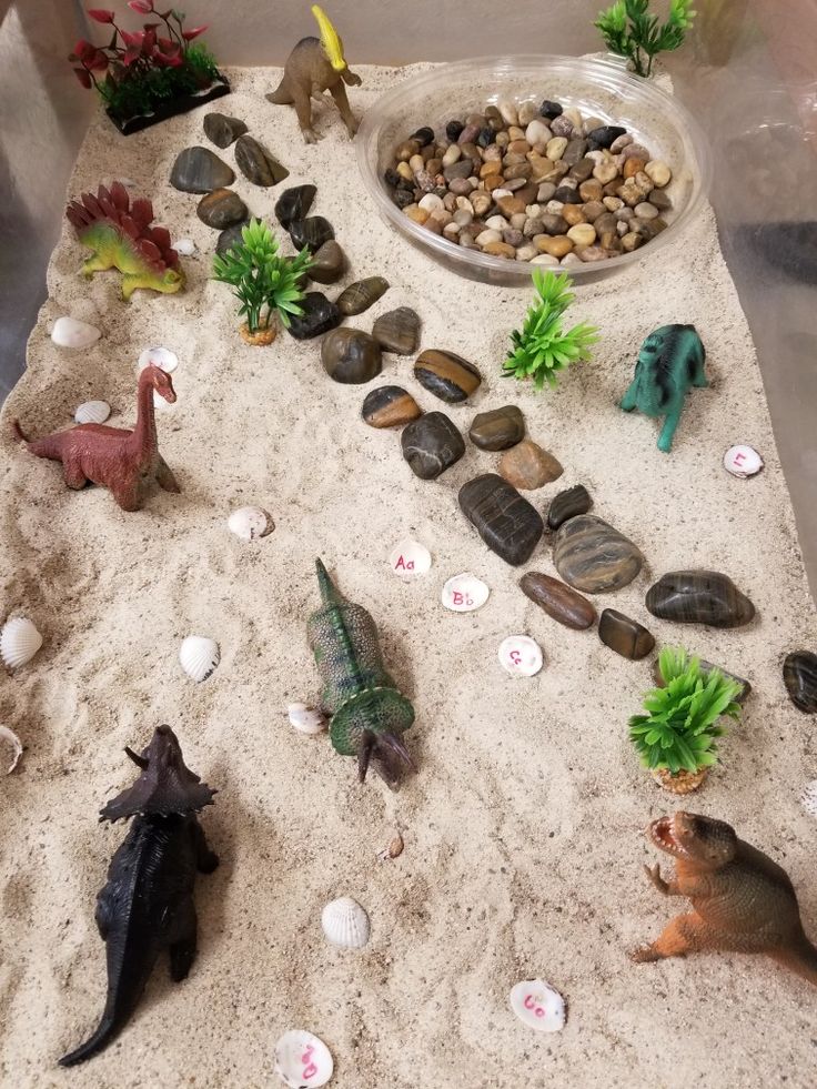 there are many toy dinosaurs in the sand with rocks and gravel on it, including one dinosaur