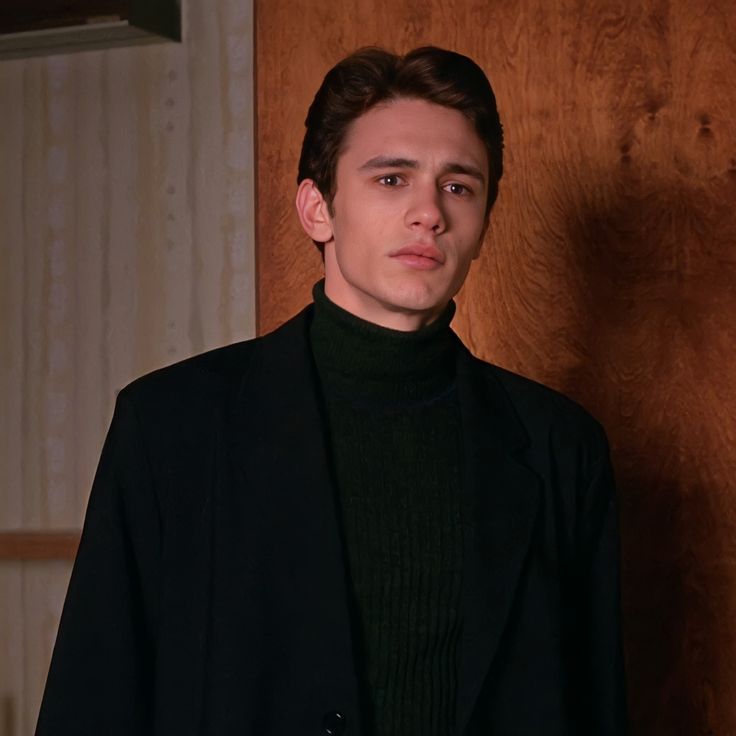 a man standing in front of a wooden wall wearing a black suit and turtle neck sweater