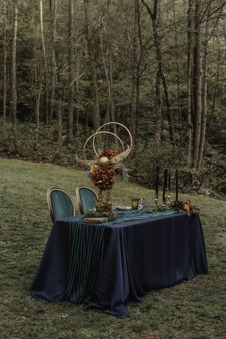 an outdoor table set up with blue cloths and gold vases on it, surrounded by trees