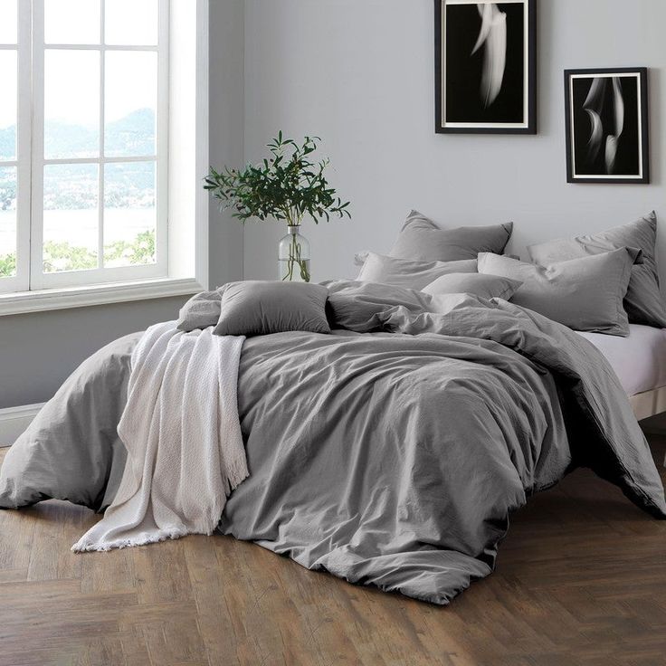 an unmade bed with grey sheets and pillows in a white room next to a potted plant