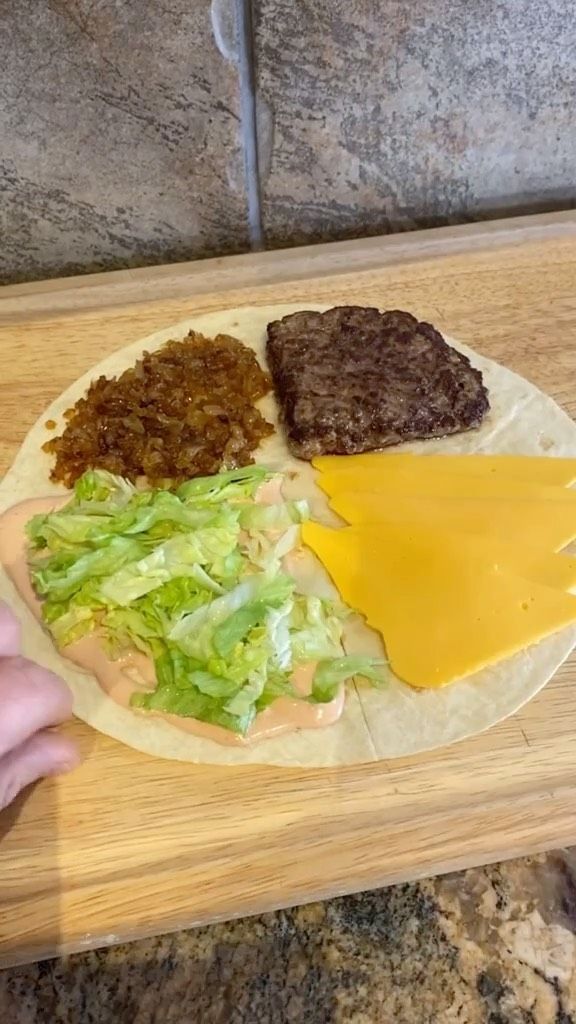 a wooden cutting board topped with cheese and meat covered in lettuce next to an uncooked hamburger