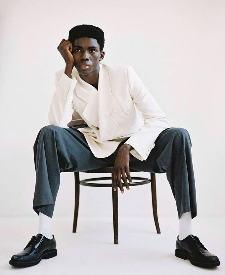 a man sitting on top of a chair wearing a white jacket and black shoes with his hand under his chin